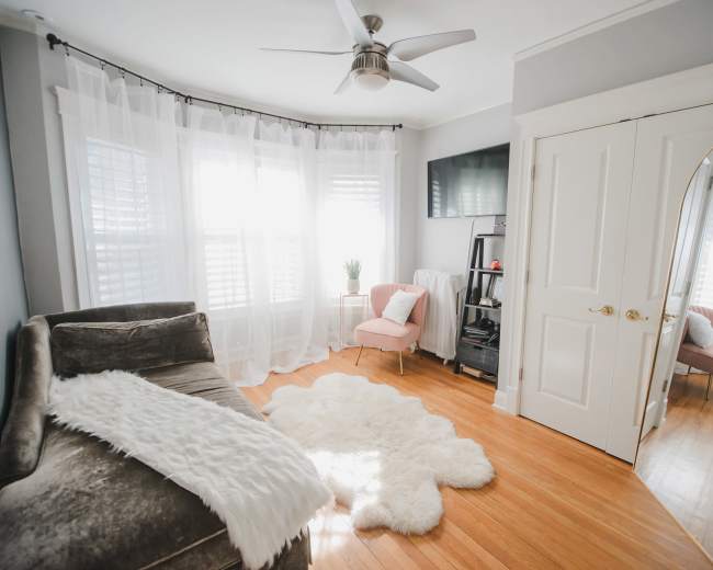 The image shows a well-lit room featuring a plush sofa, a light-colored rug, a small pink chair, and large windows dressed with sheer curtains.