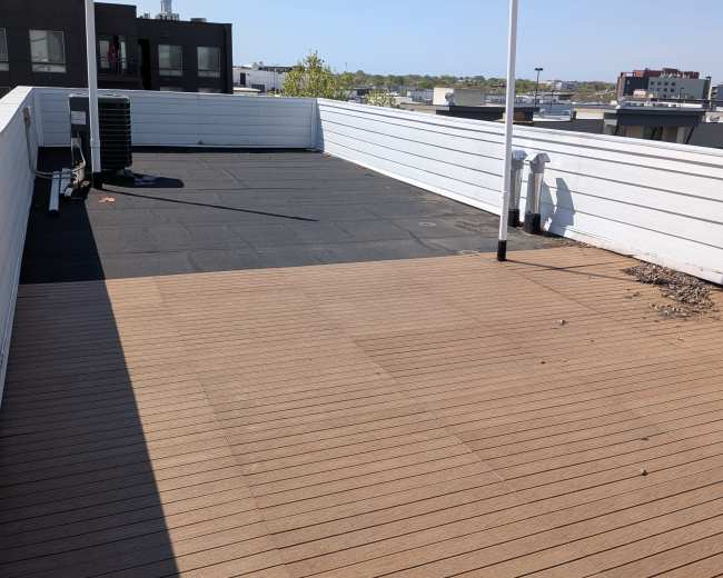 The image shows a rooftop terrace featuring a section of wooden decking and a black surface with a clear blue sky in the background.