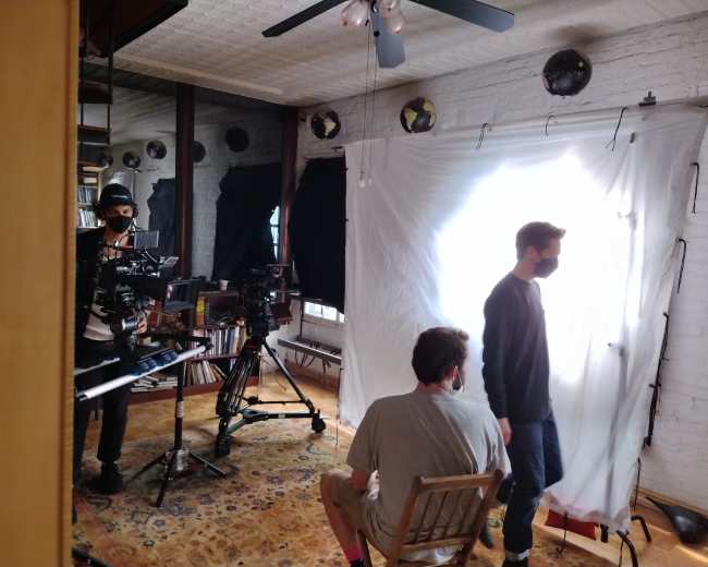 A film crew is set up in an indoor space, with one person seated and another standing in front of a bright backdrop while cameras are positioned nearby.