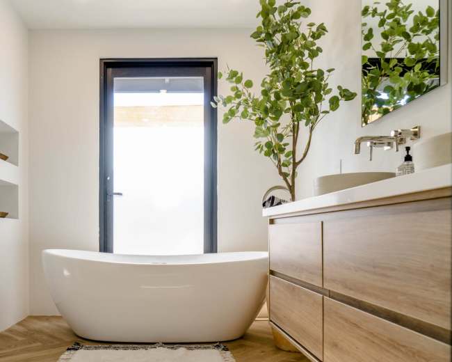 The image shows a modern bathroom featuring a freestanding white bathtub, a wooden vanity with drawers, and a large window providing natural light.