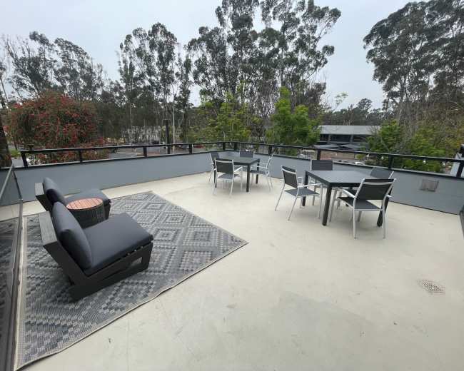 A rooftop patio featuring a seating area with a couch, a rug, and a dining set surrounded by trees.