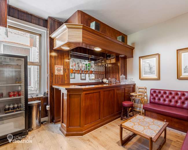 The image shows a small, wooden bar area with a red leather couch, a coffee table, and framed artwork on the walls.