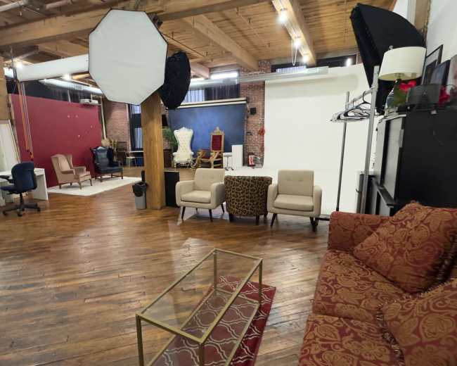 The image shows a spacious photography studio with wooden floors, comfortable seating areas, and various backdrops set up for shooting.