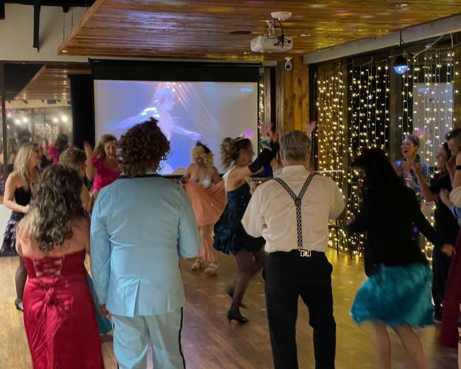 A lively group of people is dancing on a wooden floor in a decorated venue with twinkling lights and a projected image in the background.