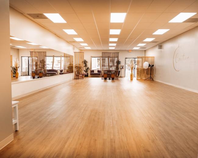 The image shows a spacious, well-lit studio with wood flooring, large mirrors, and seating areas separated by wooden dividers.