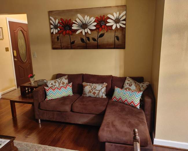 The image shows a cozy living room with a brown sectional sofa adorned with colorful patterned pillows, next to a wooden coffee table and a wall decorated with a painting of flowers.