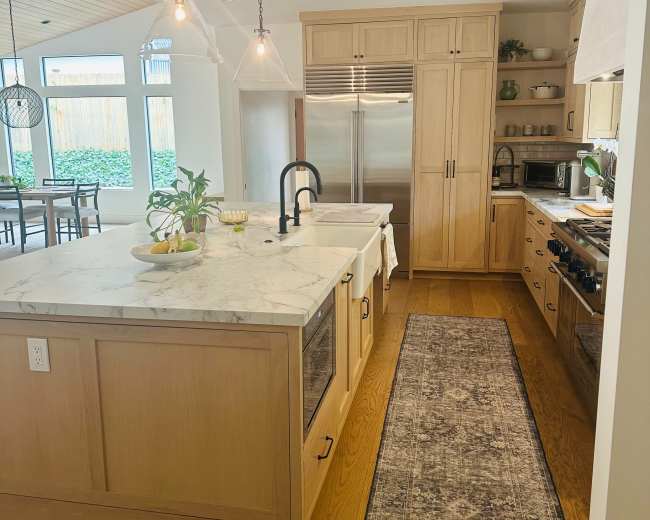 The image shows a modern kitchen featuring a large island with a marble countertop, wooden cabinetry, and stainless steel appliances, illuminated by pendant lights.