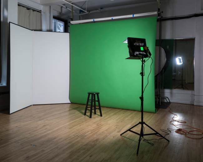 The image shows a photography studio set up with a green backdrop, a black stool, and lighting equipment.