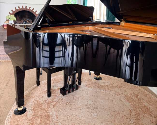 The image shows a grand piano positioned in a bright room with a fireplace and large windows.