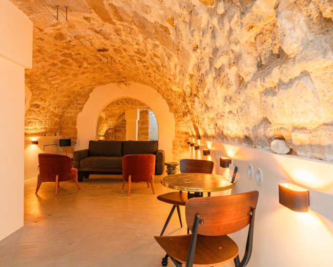 The image shows a cozy interior space featuring a stone arch ceiling, a sofa, a small round table, and two chairs, illuminated by warm wall sconces.