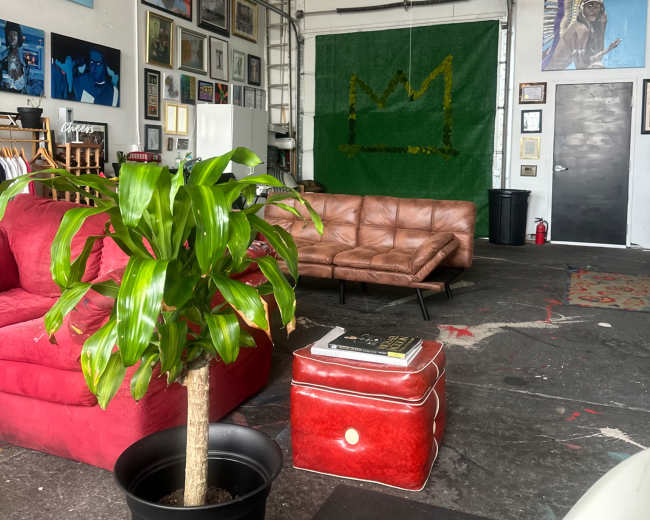 The image shows an industrial-style interior with a brown leather sofa, a red armchair, a potted plant, and various artworks on the walls.