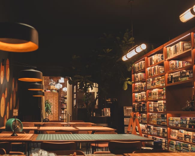 The image shows a cozy board game café with a wall decorated in vibrant circles, shelves filled with games, and wooden tables under soft lighting.