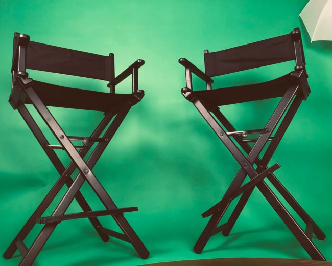 Two black folding director's chairs are positioned in front of a green backdrop.