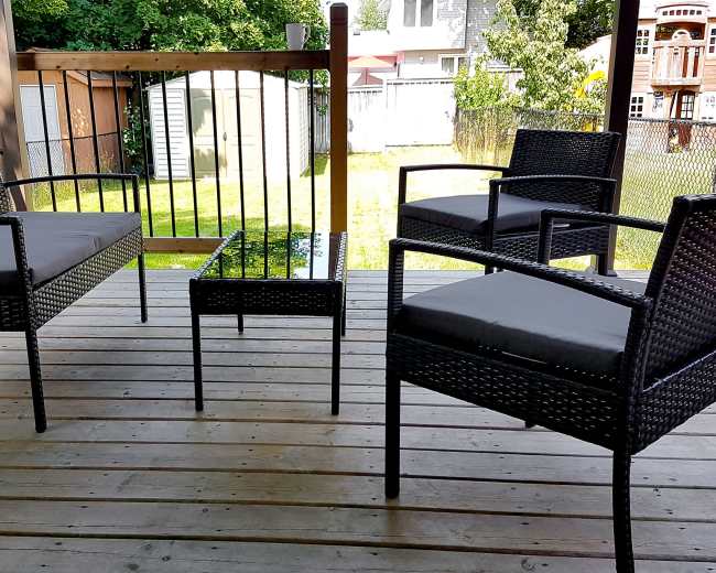 The image shows a patio with a set of black wicker chairs and a small table arranged on a wooden deck.