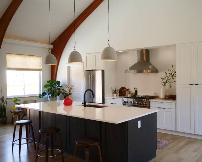 The image shows a modern kitchen featuring a large island with bar stools, stainless steel appliances, and pendant lights hanging from a high ceiling with wooden beams.