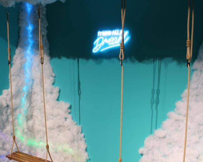 The image features two wooden swings hanging from a cloud-like backdrop with a neon sign that reads "IT'S ALL A DREAM."