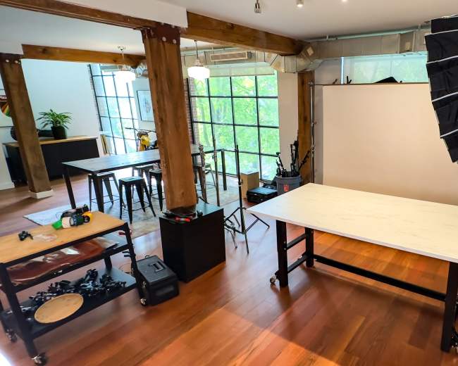 The image shows a well-lit studio space featuring a table, photography equipment, and a large backdrop setup with large windows in the background.