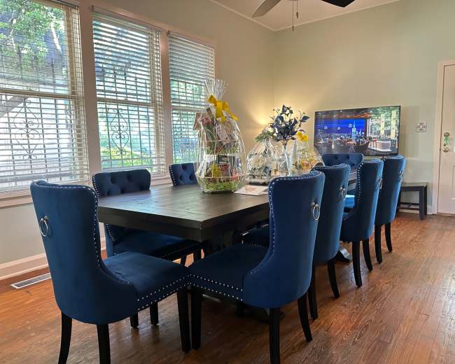 A dining room features a large wooden table surrounded by eight blue chairs, with gift baskets displayed on the table and large windows letting in natural light.