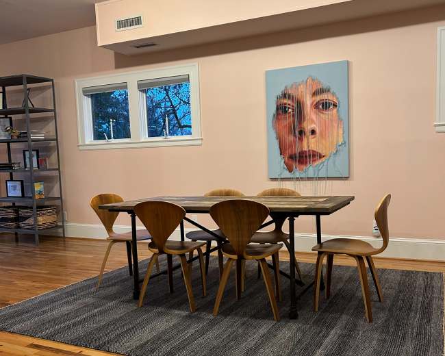 A modern dining area features a wooden table surrounded by six chairs, with a large art piece depicting a face hanging on the wall.