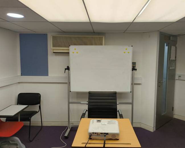 The image shows a small meeting room with a whiteboard on a stand, a projector on a table, and a black office chair facing the board.