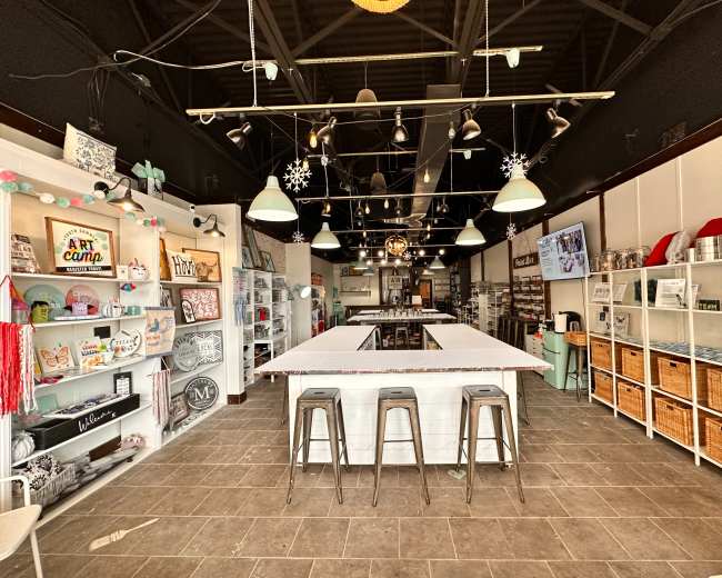 The image shows a craft store interior featuring a central work table, shelving with various art supplies, and a light fixture with pendant lamps.