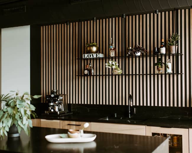 The image features a modern kitchen with a black countertop, wooden paneling on the wall, and shelves displaying various decorative items and bottles.