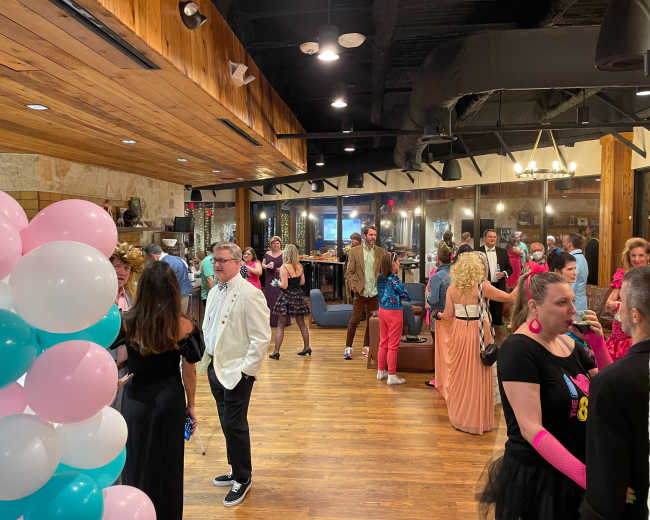 A lively indoor party scene features guests in varied attire, including formal and retro outfits, mingling around colorful balloons and decorations.