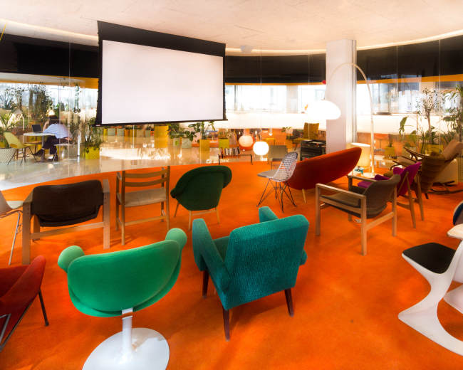 The image shows a modern lounge area with colorful chairs arranged in a semicircle facing a projection screen in a well-lit space.