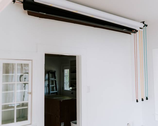 The image shows a room with a white wall featuring a roller system for backdrops mounted above a door, with colorful pulleys extending downward.
