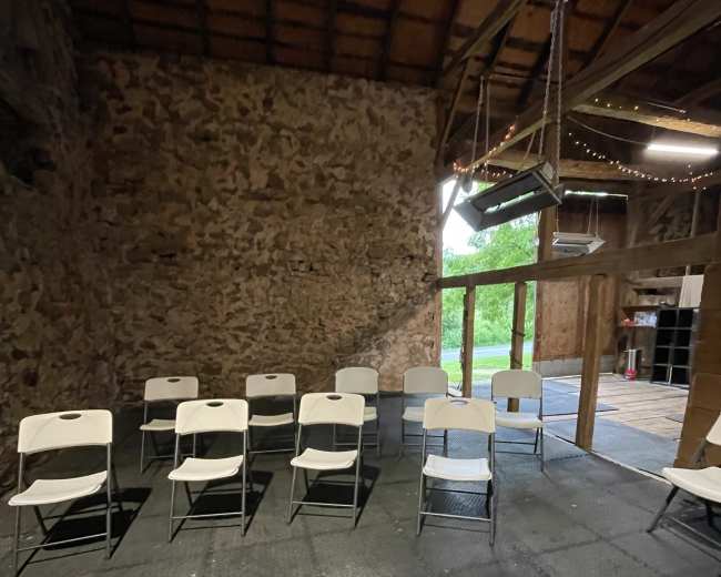 A row of white folding chairs is arranged on a textured black floor inside a rustic building with stone walls and wooden beams.
