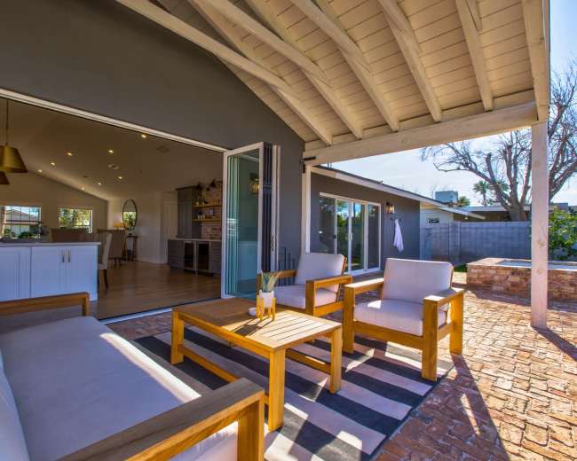 The image shows a modern outdoor seating area with wooden furniture and an open view of a sunlit interior space.