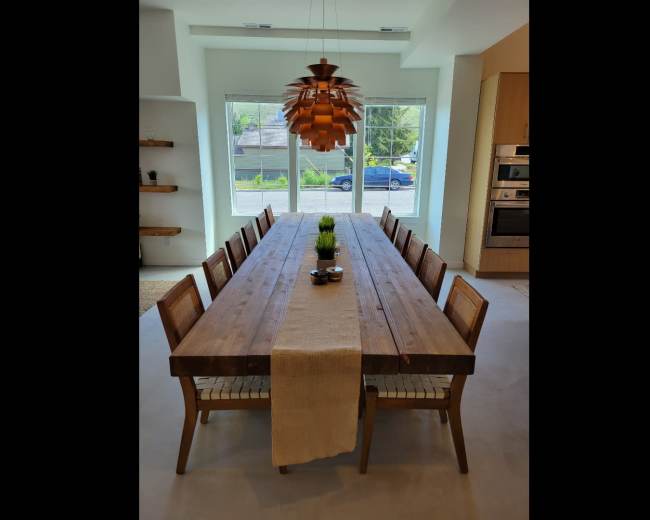 A long wooden dining table with eight chairs is centered under a pendant light, facing large windows that overlook a street.
