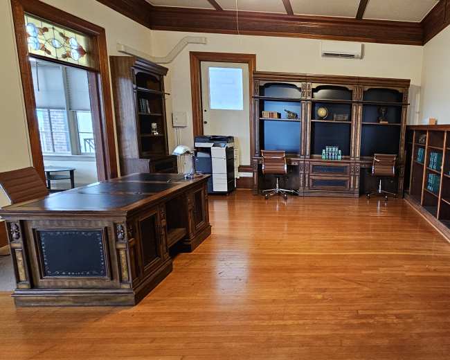 A spacious office with wooden flooring, a large dark wooden desk, shelves filled with books, and a doorway leading to another room.
