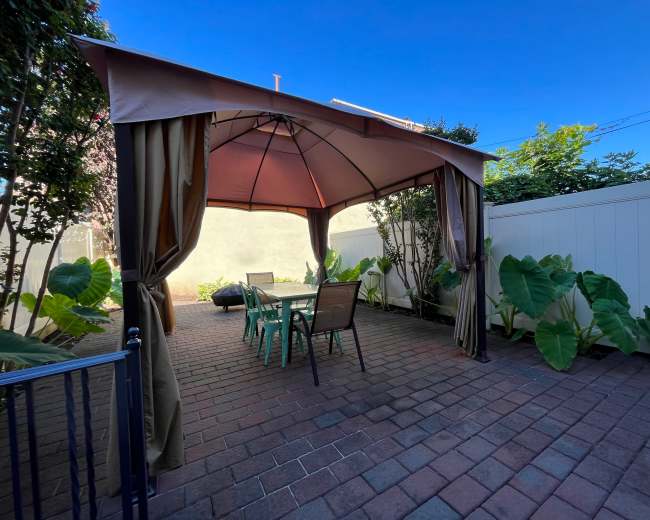 A gazebo with draped fabric encloses a dining table and chairs, surrounded by large green plants in a brick-paved courtyard.