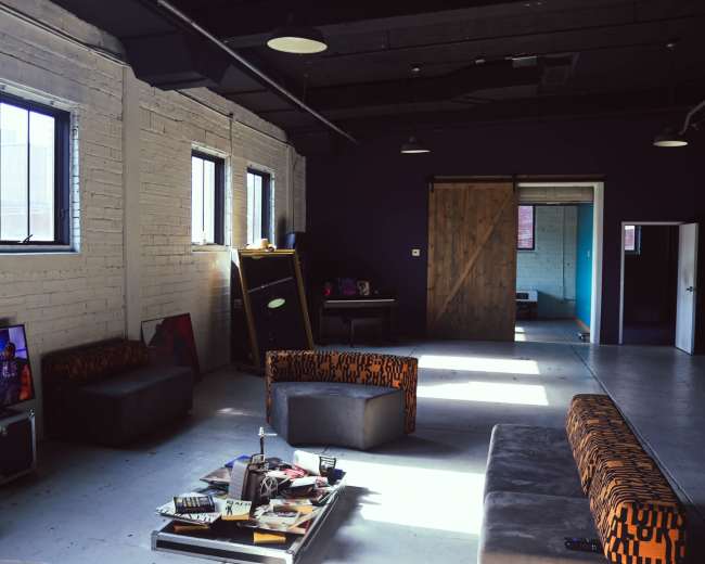 The interior of a spacious room features two large windows, a collection of low sofas with orange and black patterns, a coffee table with scattered items, and an open doorway leading to another area.