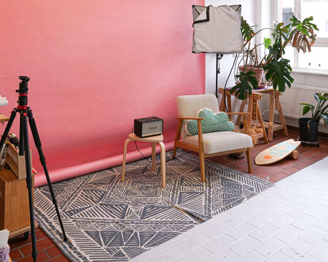 The image shows a bright studio interior featuring a pink backdrop, a patterned rug, a wooden chair, and a camera setup.