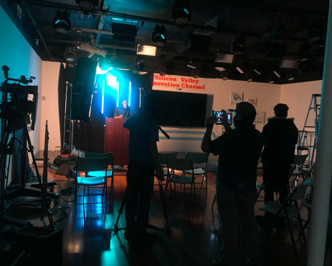 A person stands at a podium in a studio setting, while cameras and a small audience are positioned in front of them.