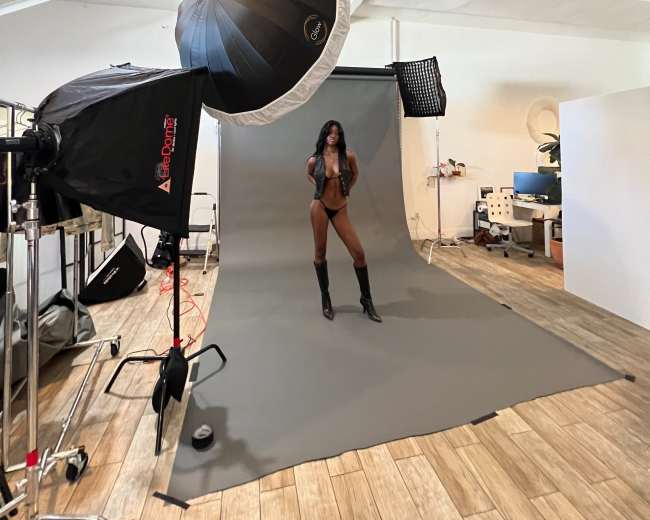 A model poses on a gray backdrop in a photography studio equipped with lighting and camera equipment.