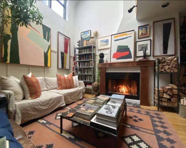 The image depicts a cozy living room with a sofa, a bookshelf filled with books, a fireplace with a fire burning, and artwork hanging on the walls.