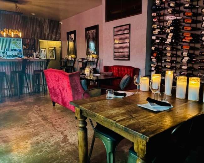 The image shows a dimly lit restaurant interior featuring wooden tables, red upholstered seating, and a wall displaying a wine rack.
