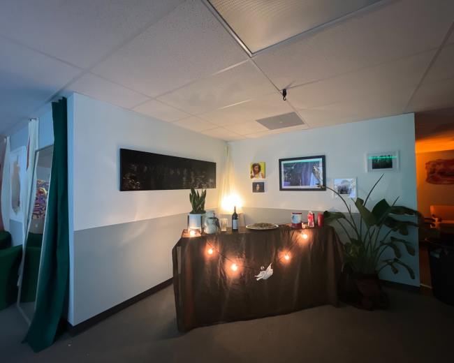 A corner of a room features a table draped in dark fabric, adorned with a lamp, plants, and various photographs, with string lights positioned around the display.