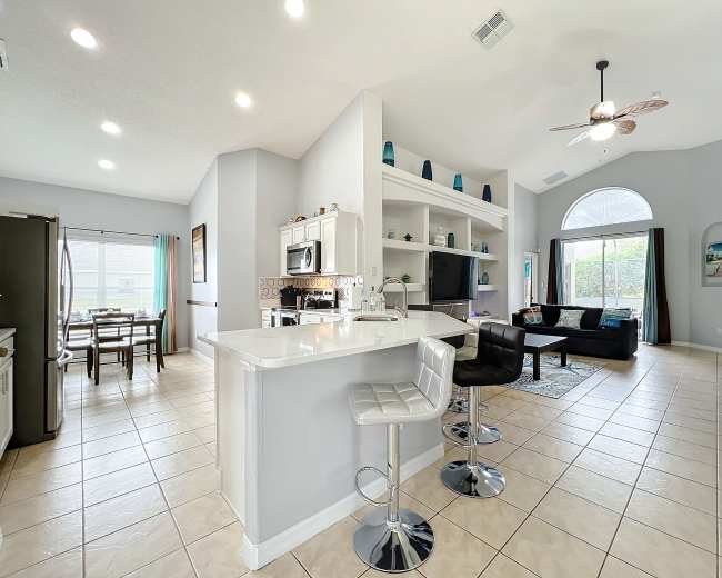 The image shows a spacious open-concept living area and kitchen, featuring a breakfast bar, dining space, and a large window with a comfortable seating area.