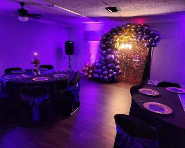 A decorated room features wooden flooring with two tables set with plates and a large arch of balloons in purple and gold against a backdrop of fairy lights.