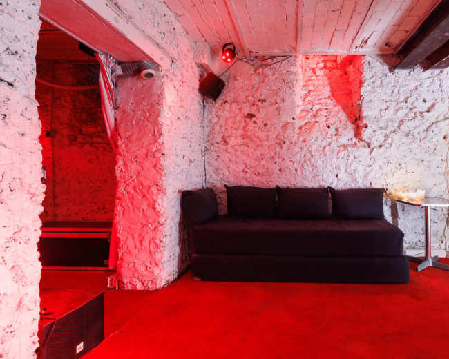A small, dimly lit room with stone walls features a dark couch and a side table on red carpeting.