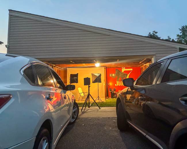 Two vehicles are parked in front of a garage, which has a brightly lit interior featuring a red wall and a chair set up for an event or shoot.