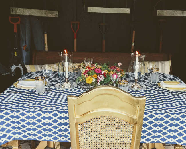A dining table is set with a blue and white patterned tablecloth, candles, flowers, and glassware, surrounded by gardening tools hanging on the wall behind it.