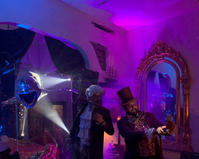 Two men in period costumes pose in a dimly lit room decorated with theatrical lighting, mirrors, and eclectic furnishings.