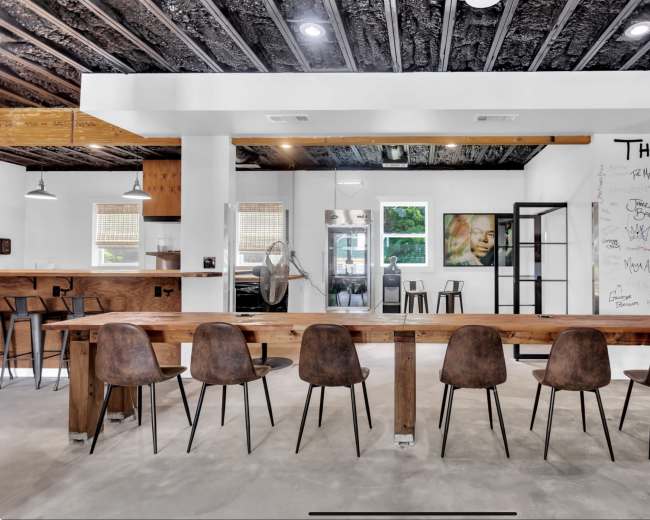 A spacious, modern coworking area features a long wooden table with several chairs, a kitchen counter, and walls adorned with drawings and handwritten notes.