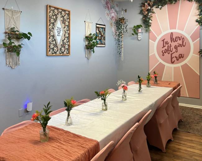 The image shows a decorated room set for a gathering, featuring a long table with floral centerpieces and wall art, along with hanging plants and star-shaped decorations.
