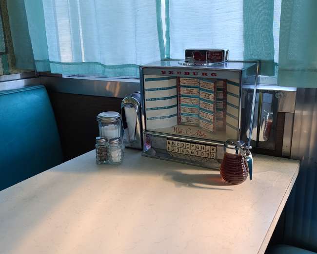 A diner table with a Seeburg jukebox, salt and pepper shakers, and a glass bottle of syrup, all beneath a window with light blue curtains.